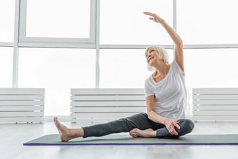 A woman in her sixties stretching on an exercise mat by a bright window